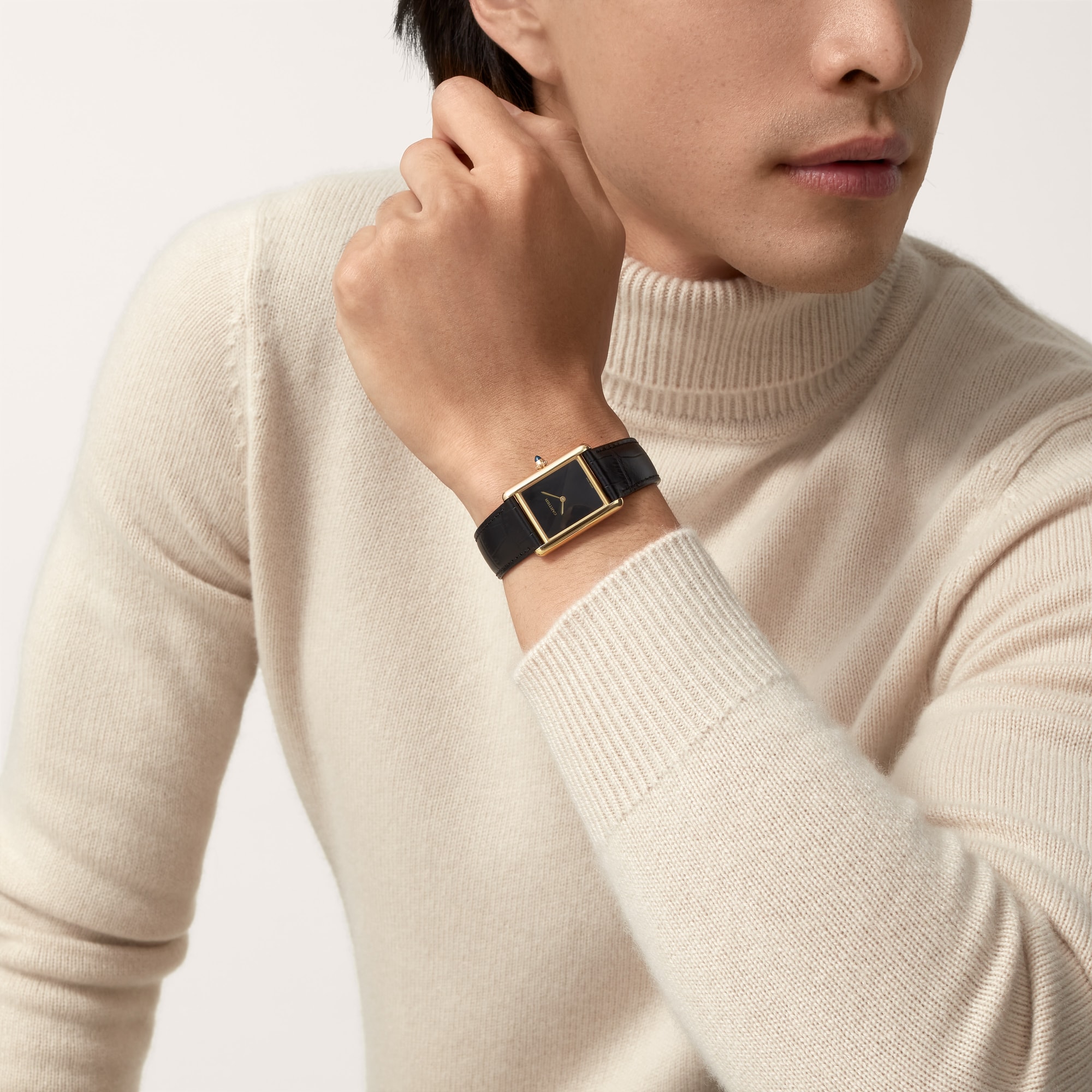 Fashionable on-wrist shot of a Dupe Tank Louis Cartier large model yellow gold color watch with a black dial and black leather strap, worn by a male model.