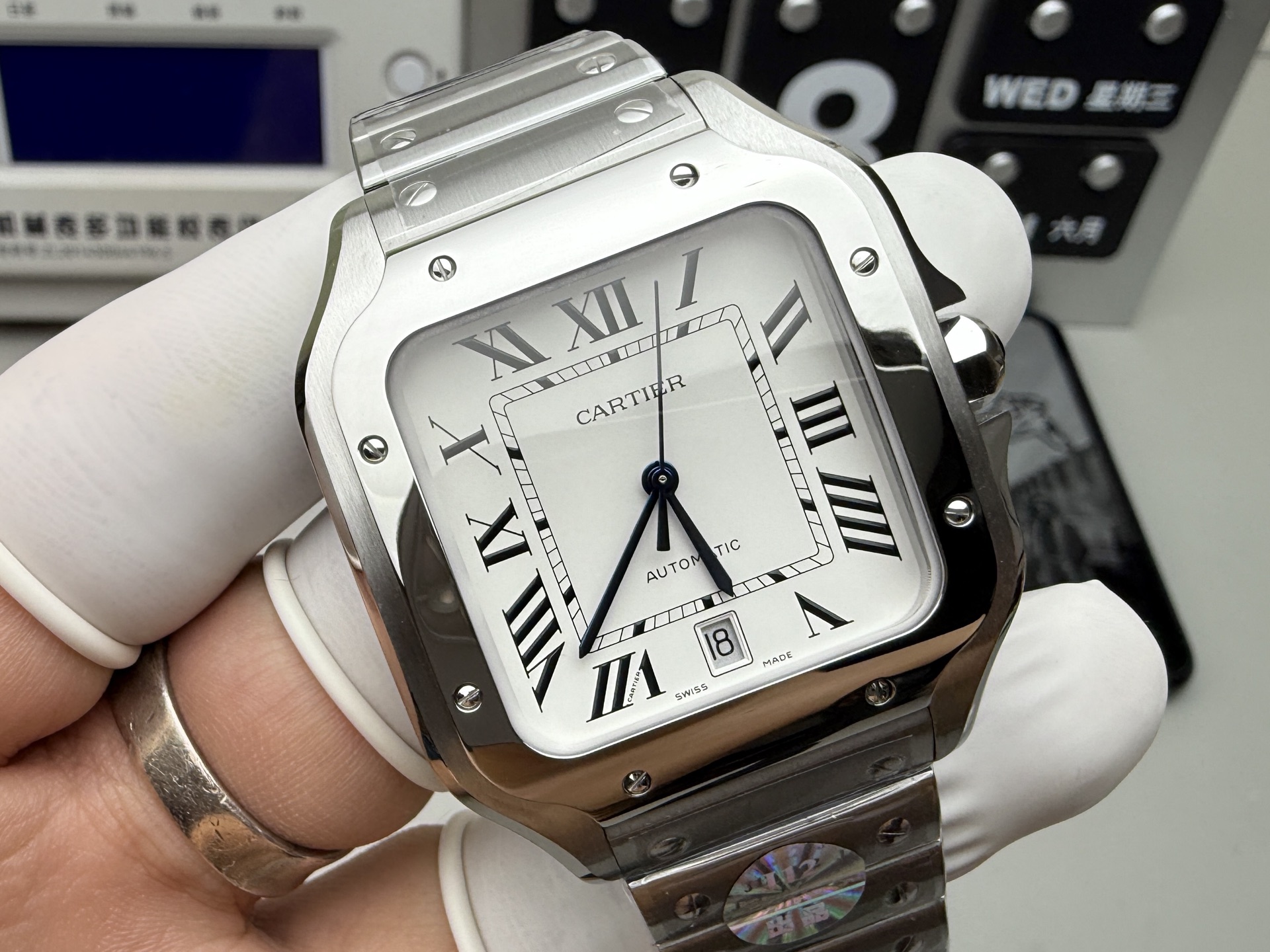 Close-up of the dial of the BVF Super Clone Cartier Santos replica watch, silver case, white dial, date display, and steel bracelet, held by a gloved hand, emphasizing Roman numerals and hands.